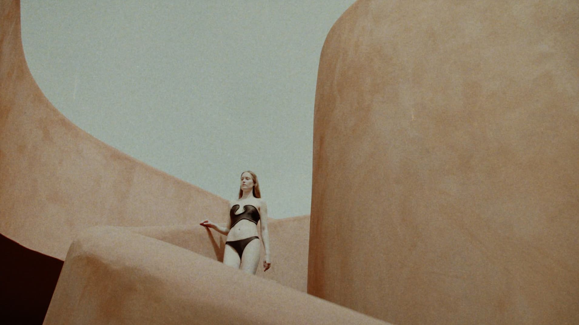 Model wearing a black two-piece swimsuit standing amidst curved architectural terracotta walls under an open sky