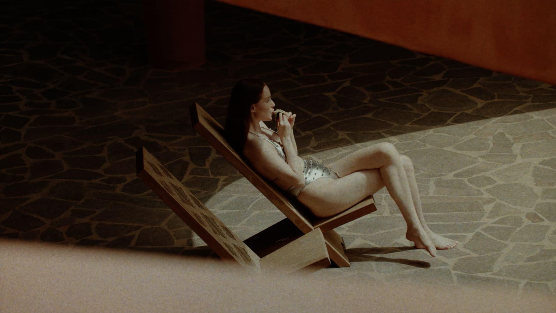 Model seated on a wooden lounge chair holding food against a stone patio background