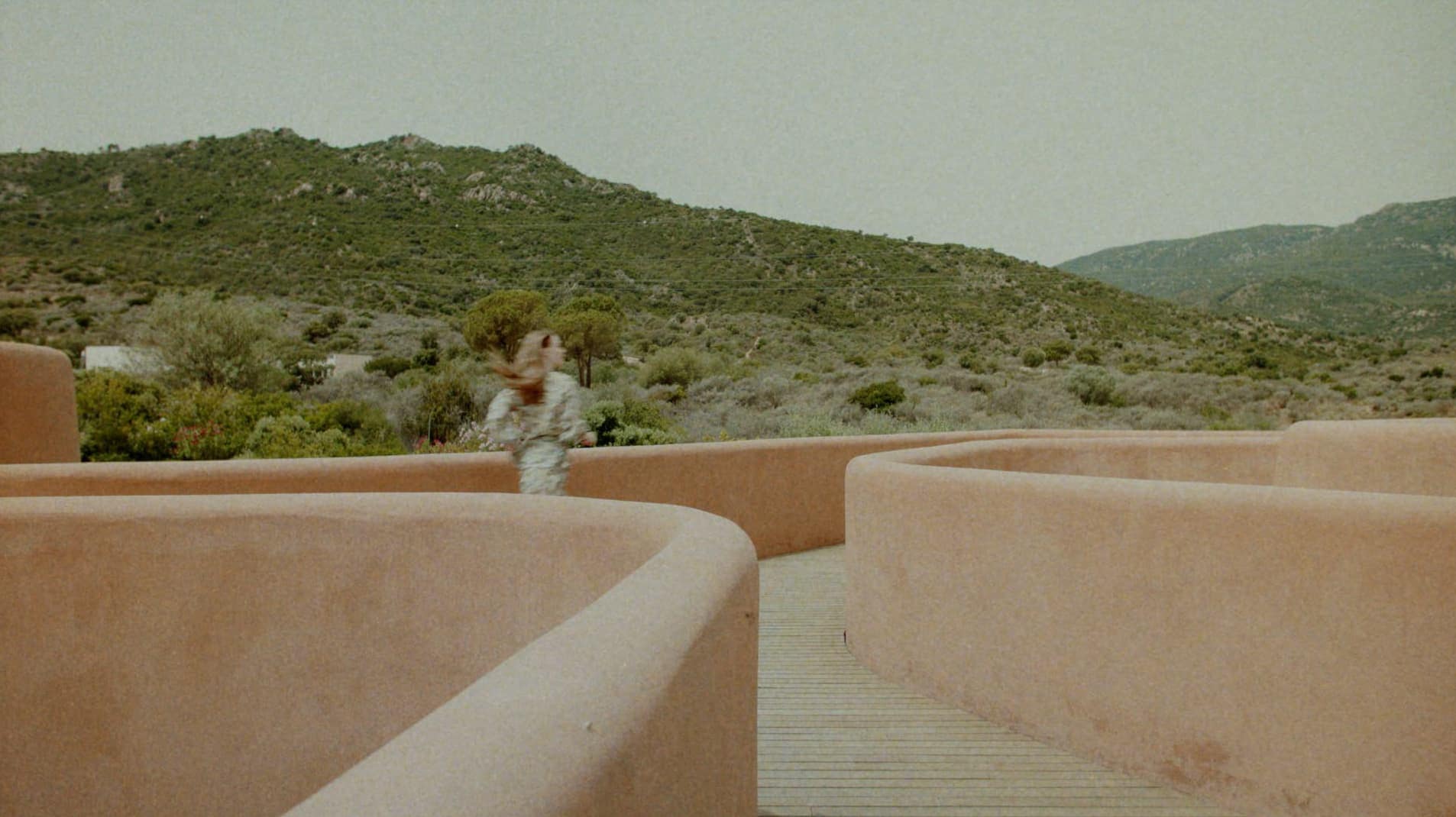 A person walking along a curved terracotta walkway with green hills and overcast sky in the background.