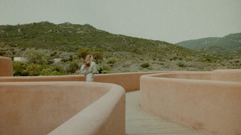 A person walking along a curved terracotta walkway with green hills and overcast sky in the background.