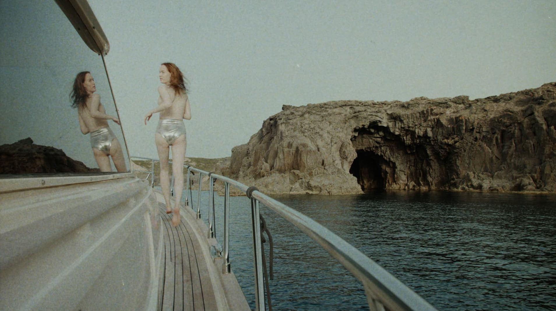 Model walking on the deck of a boat wearing silver metallic swimwear with rocky coastal cliffs visible in the background