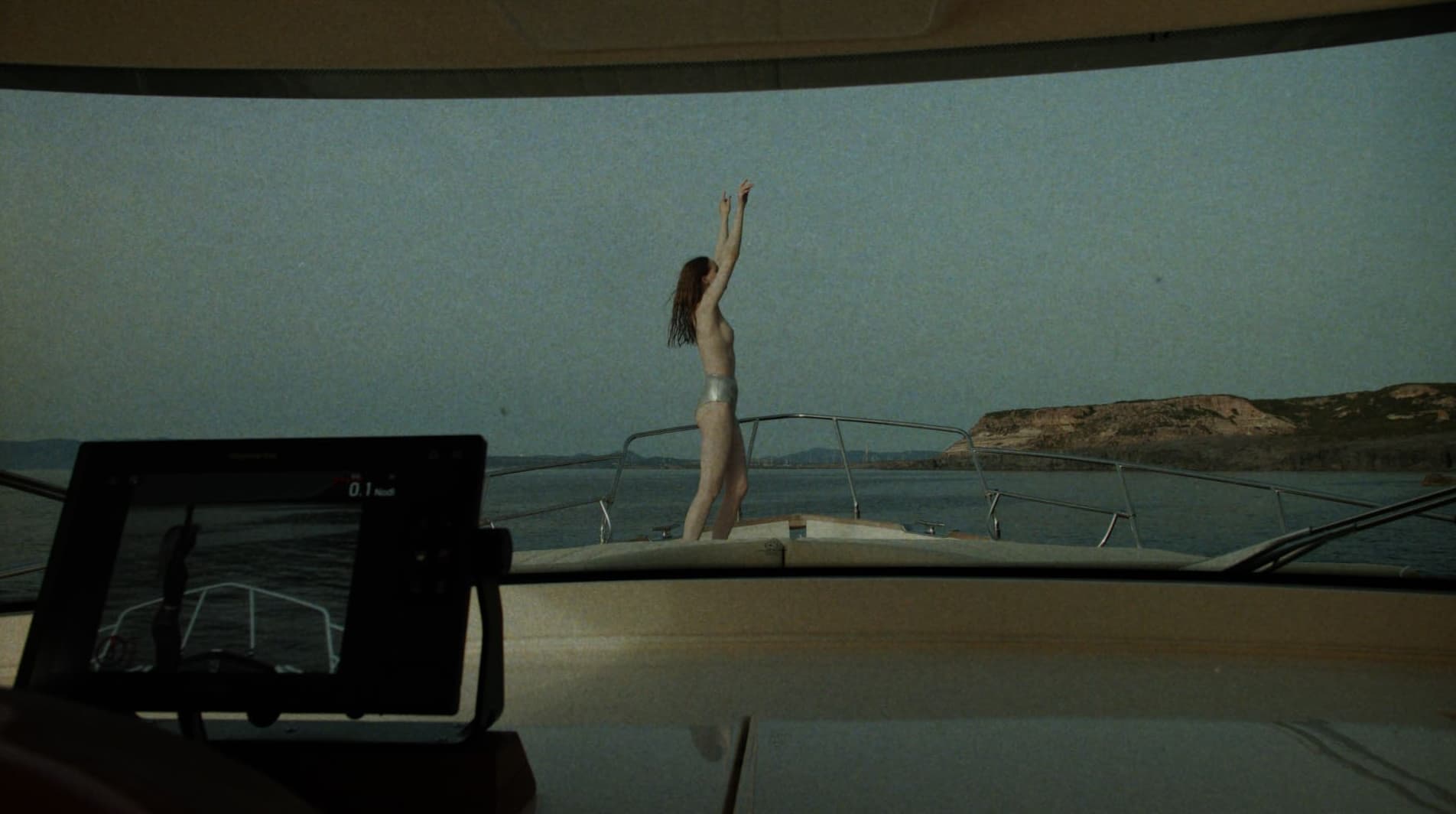 Model standing on the bow of a yacht with arms raised against an open sea and distant coast