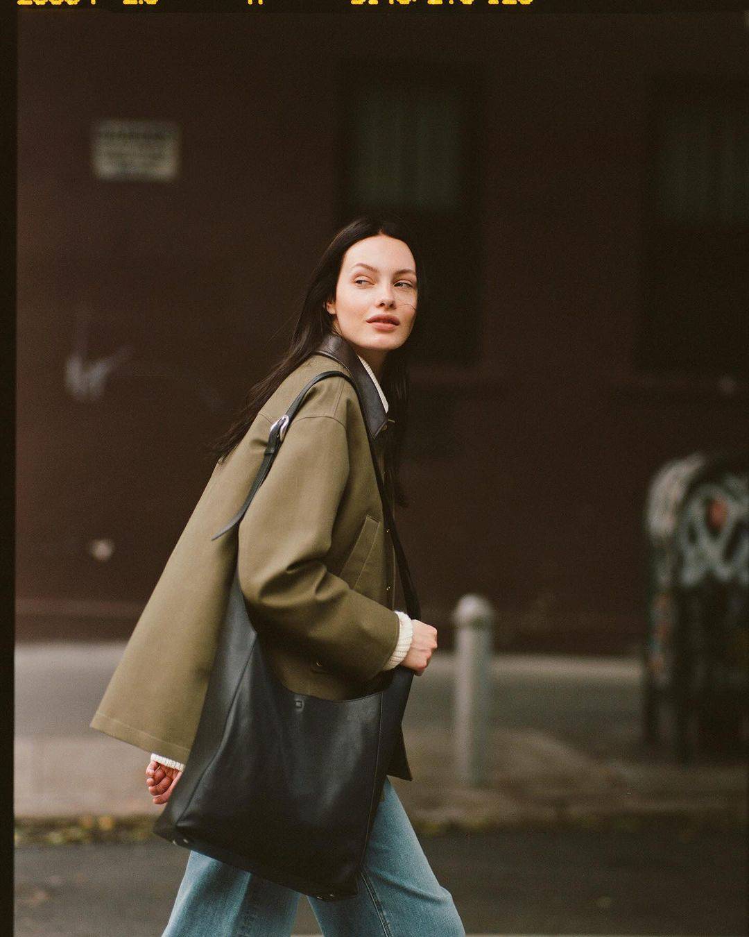 Woman in an olive green jacket and blue jeans walking down a city street while carrying a large black leather shoulder bag.