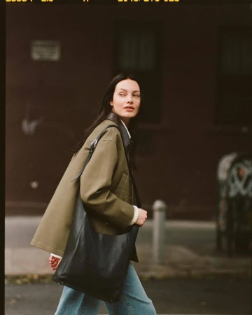 Woman in an olive green jacket and blue jeans walking down a city street while carrying a large black leather shoulder bag.