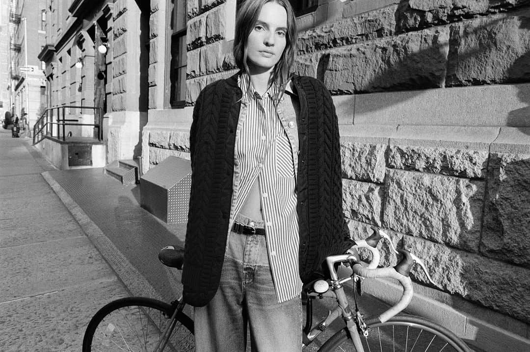 Model standing on a city sidewalk next to a bicycle in a black and white fashion campaign photograph.