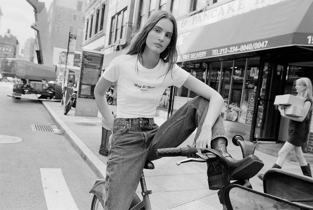 Model seated on a bicycle in the street wearing a white t-shirt and denim jeans for Rag and Bone.