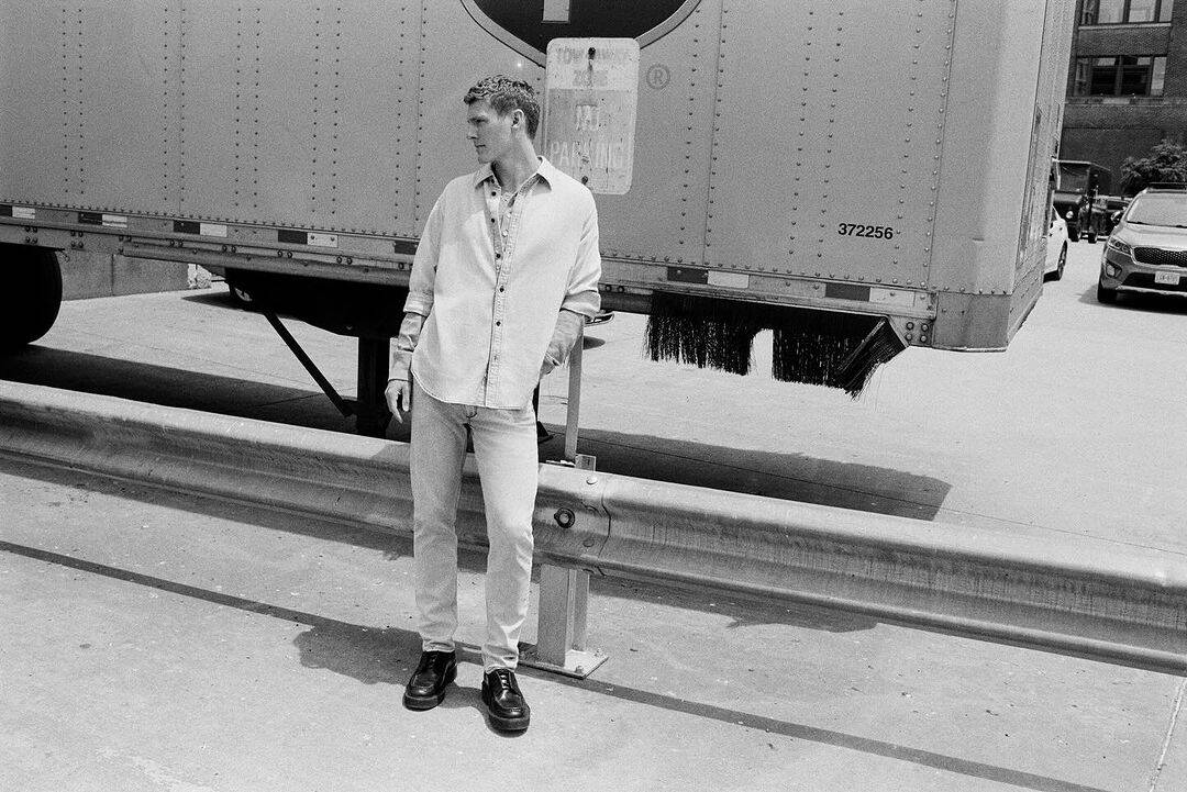 Man wearing casual button-down shirt and trousers leaning against a large truck trailer outdoors in a black and white photograph