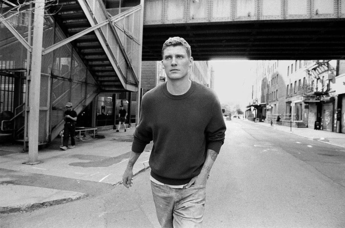 Black and white portrait of a man walking on a city street wearing a dark crewneck sweater and jeans under an elevated bridge.