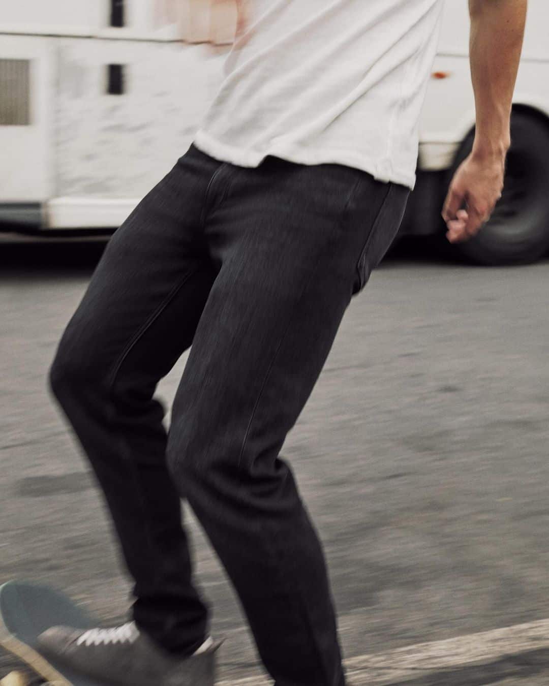 Man wearing a white t-shirt and dark jeans skateboarding outdoors on an asphalt street