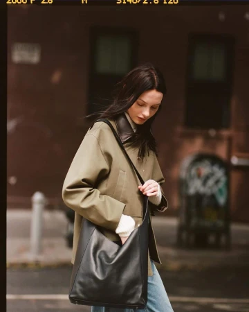 Model wearing an olive green jacket and carrying a black leather shoulder bag on a city street.