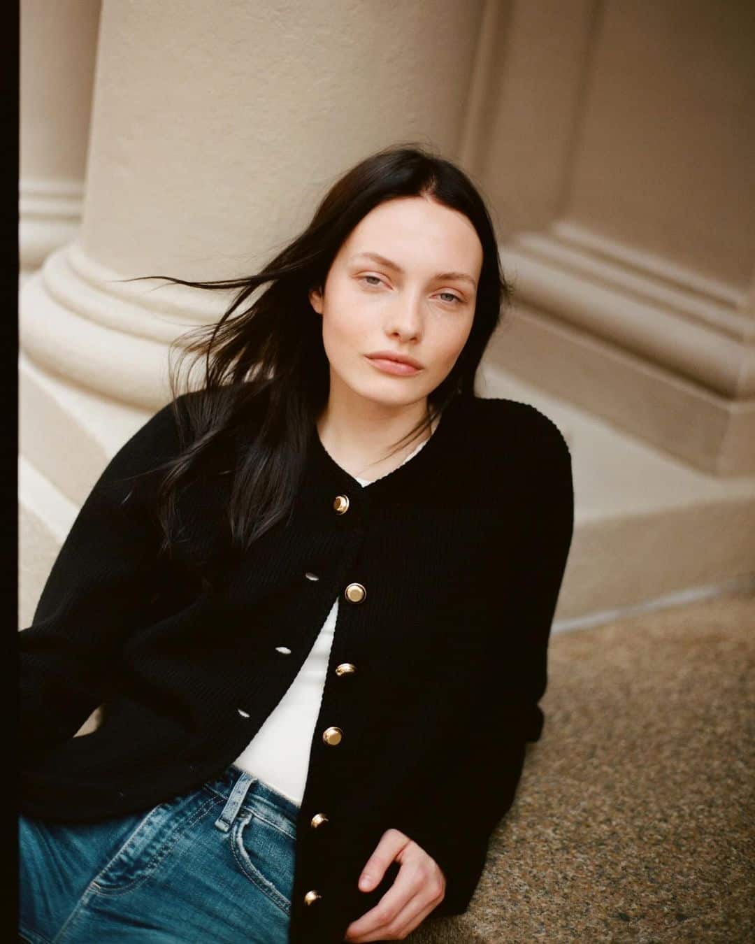 Model Tilda Lindstam wearing a black cardigan and blue jeans leaning against architectural columns