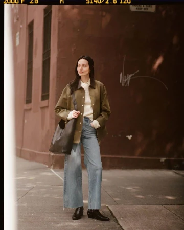 Model standing on a city sidewalk wearing an olive green jacket, white knit top, and wide-leg denim jeans carrying a dark leather shoulder bag.