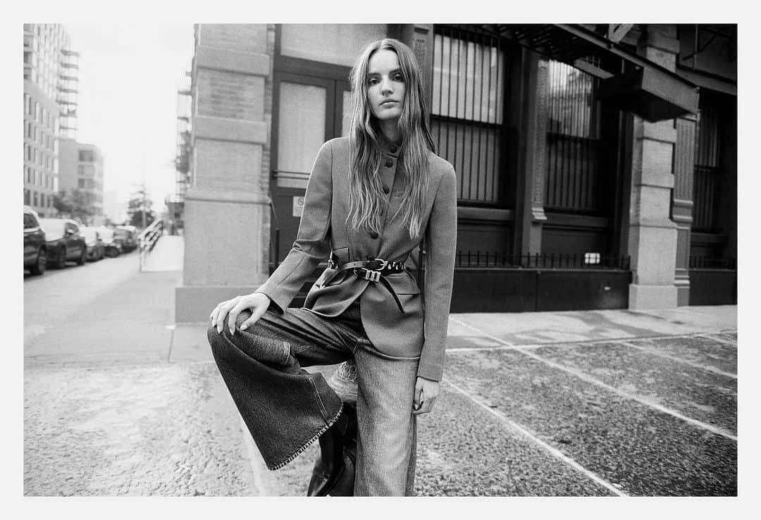 Monochrome fashion campaign image of a female model in a belted jacket and wide-leg jeans posing on an urban street.
