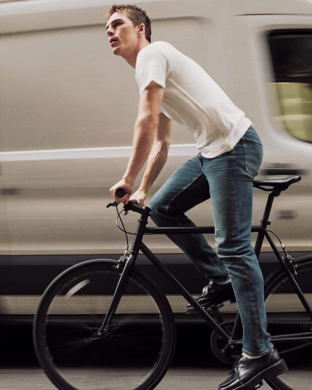 Man riding a black bicycle in front of a moving white van wearing a white t-shirt and blue jeans
