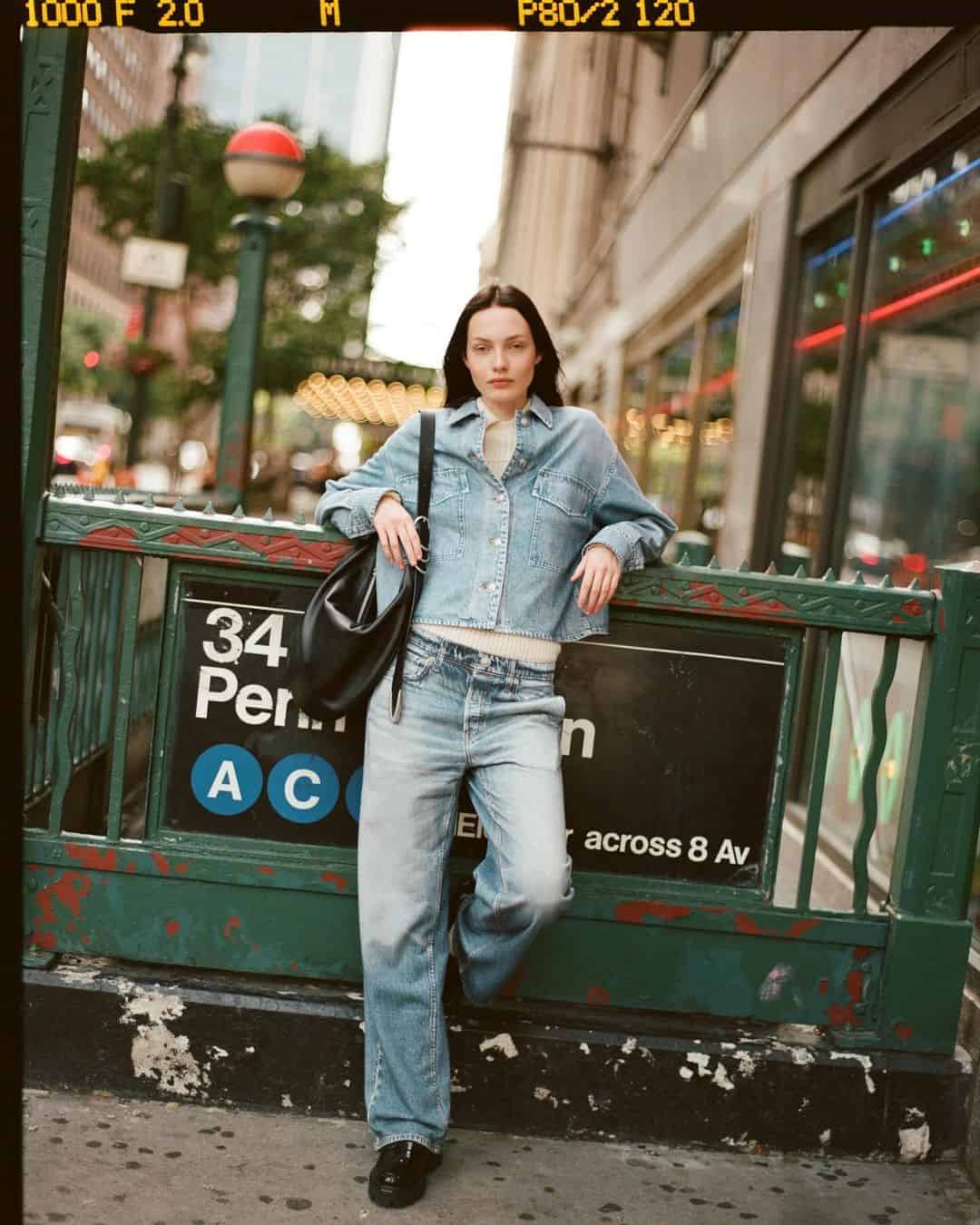 Model wearing denim shirt and jeans leaning against a green subway station entrance holding a black handbag.