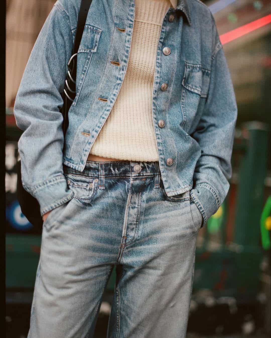 Model wearing a blue denim jacket over a cream sweater and matching jeans, photographed outdoors.