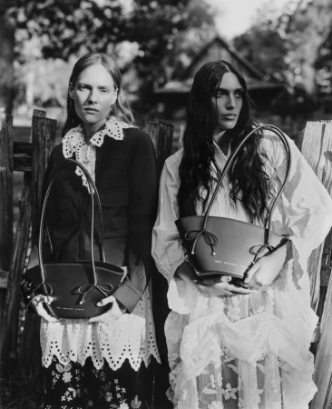 Two female models holding leather handbags outdoors in front of a wooden fence, photographed in monochrome.