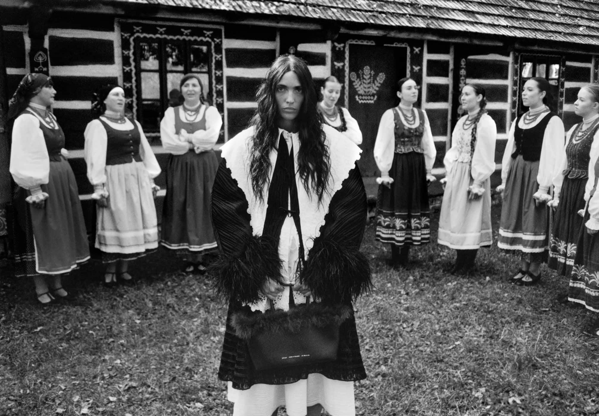 Central woman holding a dark handbag standing before a line of women in traditional folk dress outside a wooden house.