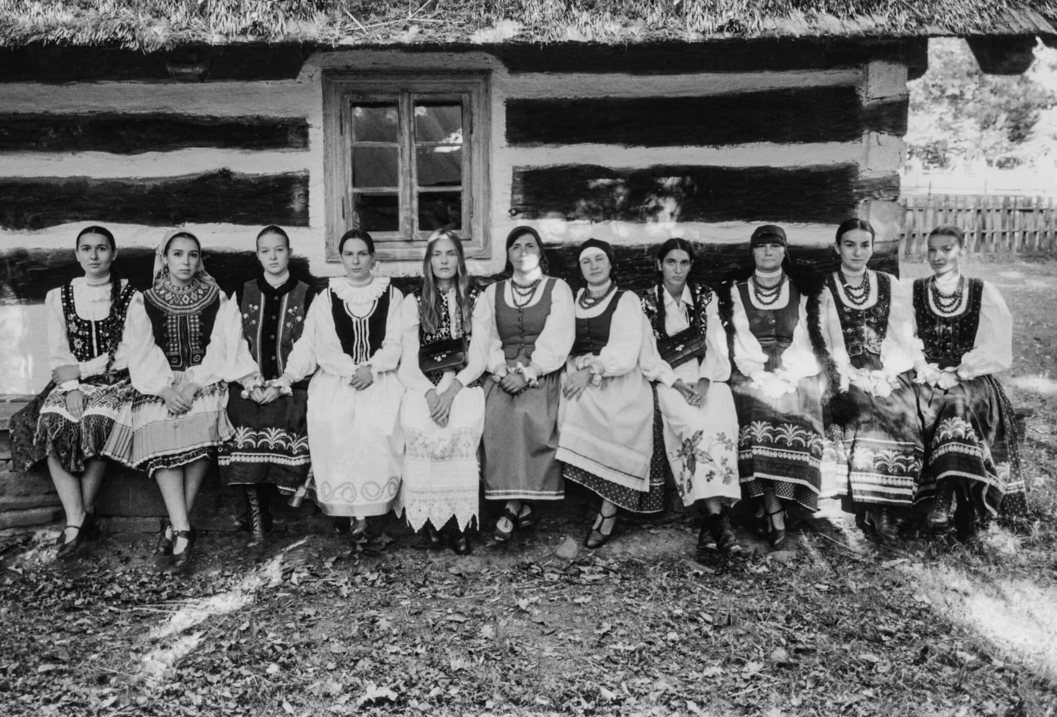 Group of women in traditional regional folk dresses seated in a row against a rustic wooden log cabin wall.