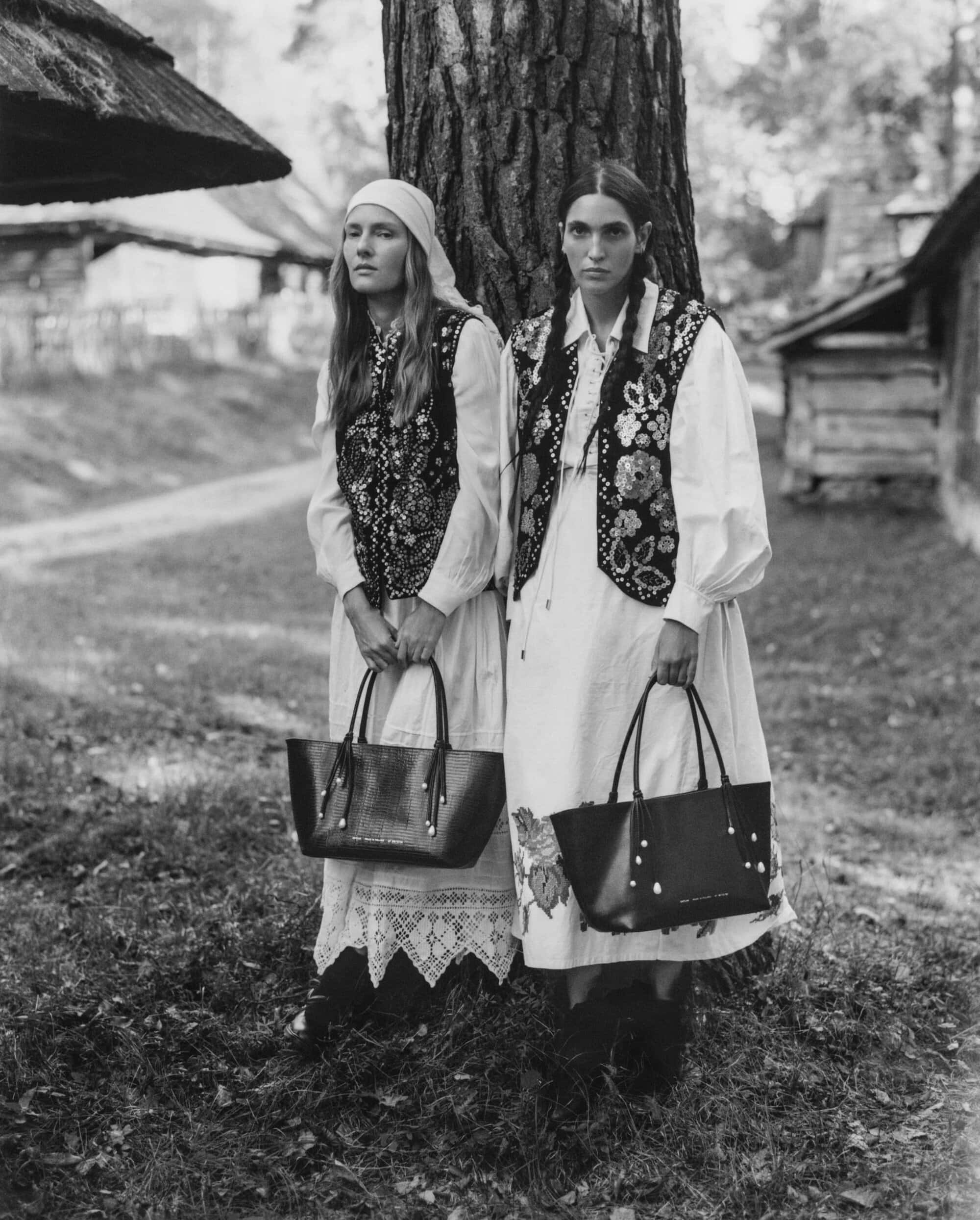 Two models in traditional white dresses and patterned vests leaning against a tree holding black leather handbags in a rural outdoor setting