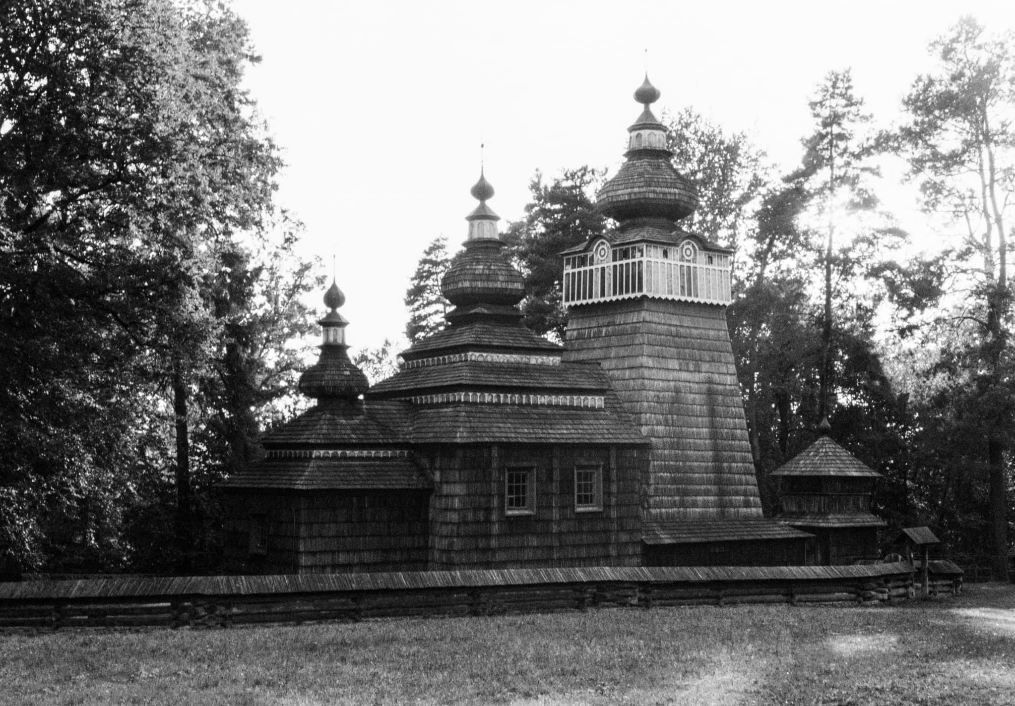 Black and white photograph of a traditional wooden church with multi-tiered cupolas surrounded by trees and a low wooden fence