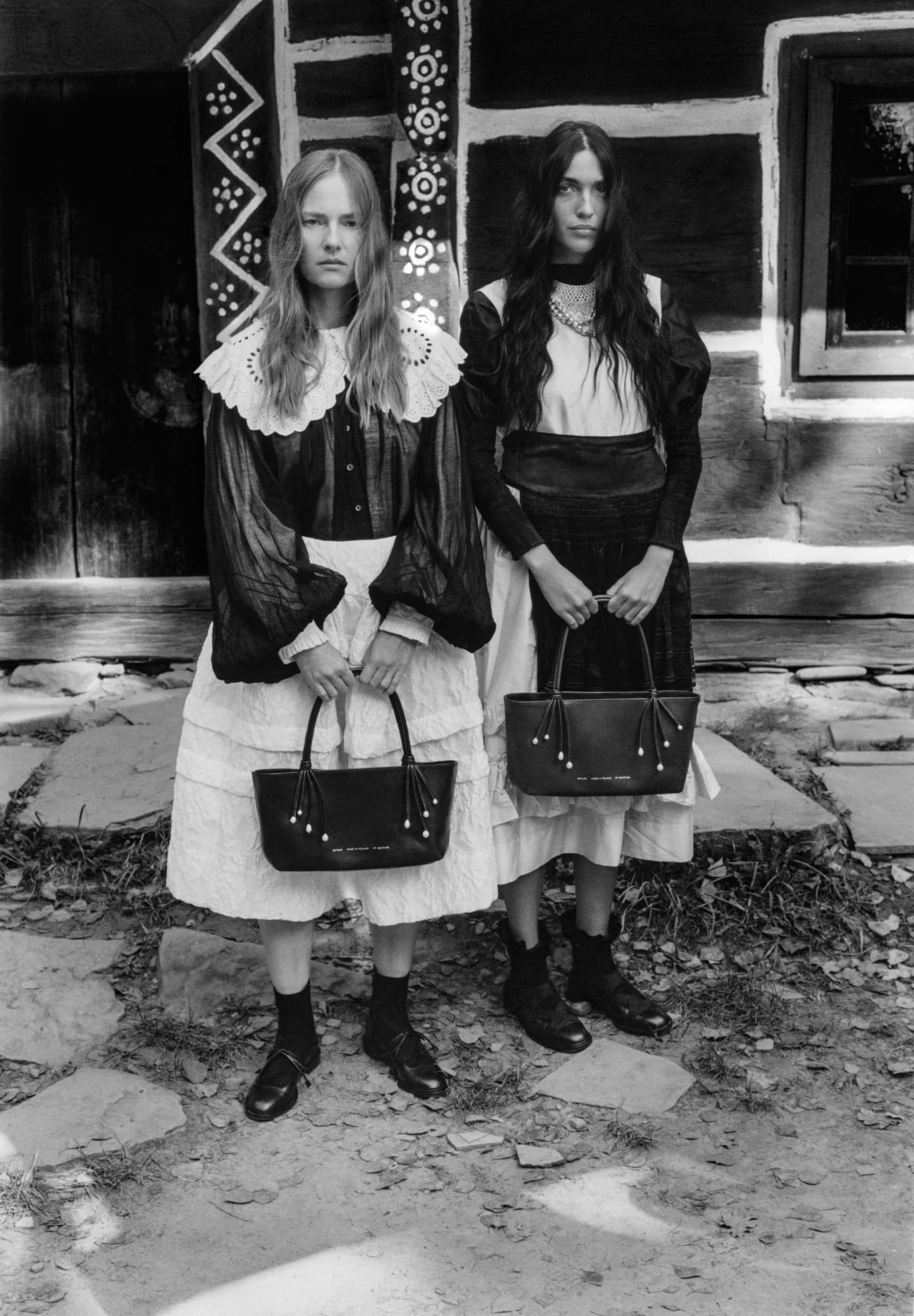 Two women posing in front of a traditional wooden building holding black leather handbags for Chylak campaign