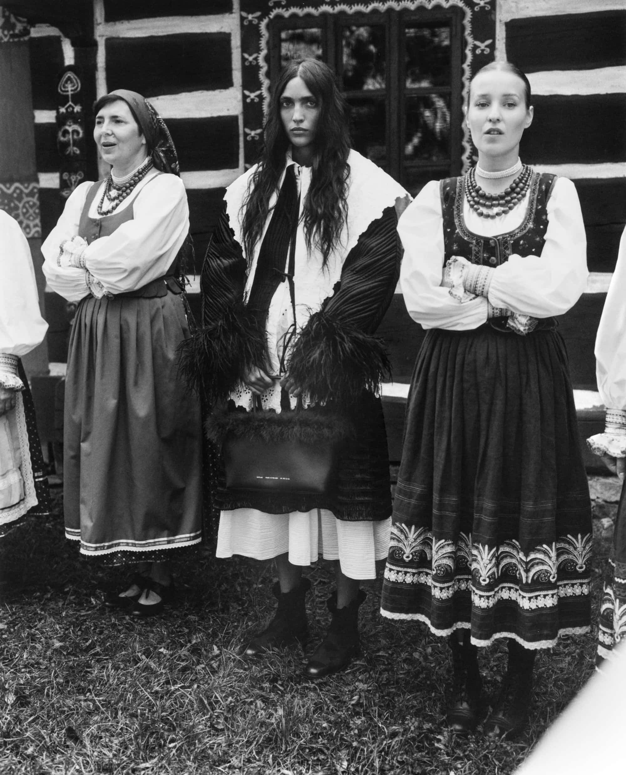 Three women in traditional folklore-inspired attire standing outdoors in front of a rustic wooden building exterior.