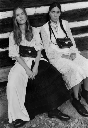 Two female models wearing white dresses and dark handbags seated against a dark rustic wooden wall.