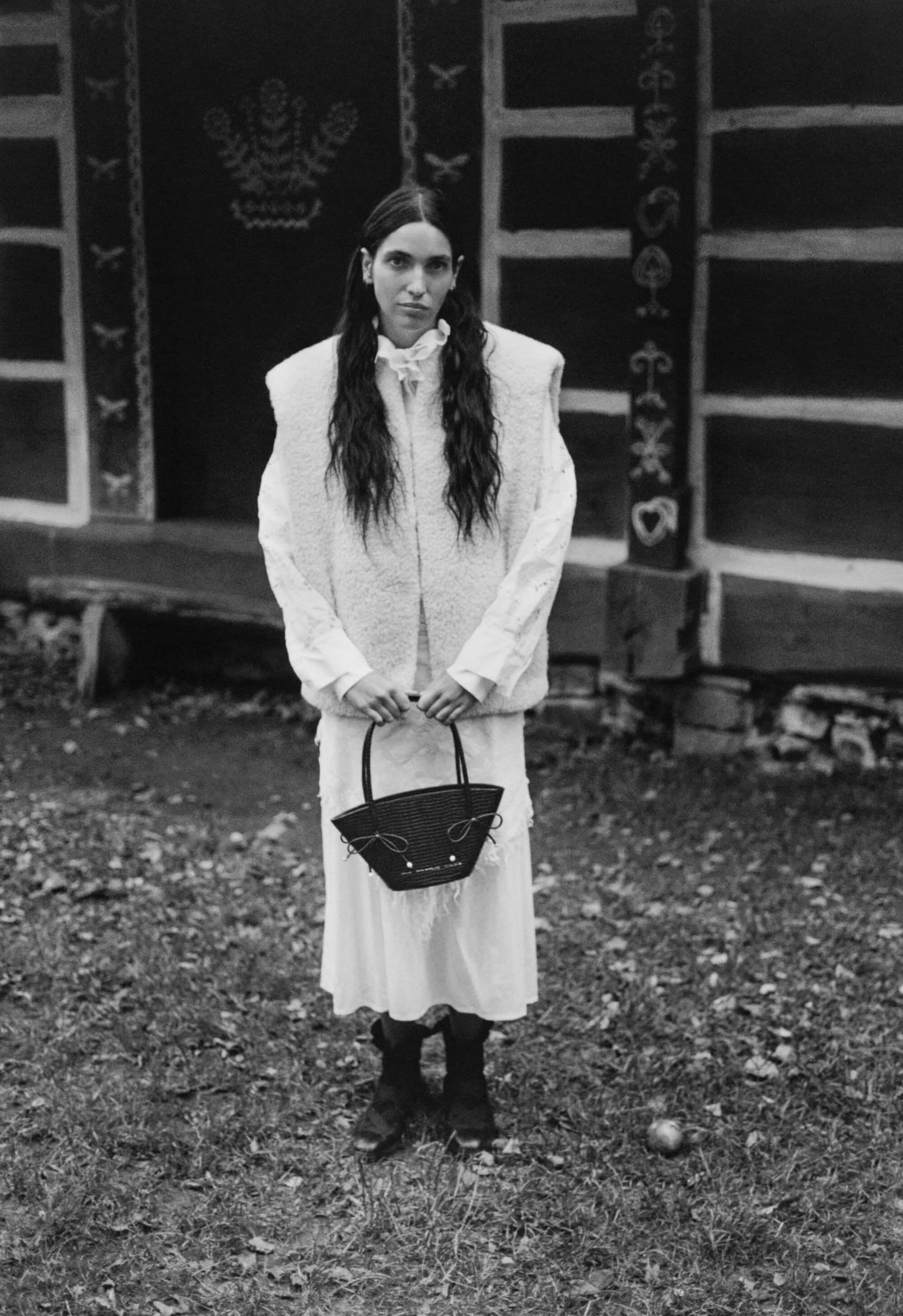 Model standing outdoors holding a dark structured handbag in front of a traditional wooden building facade