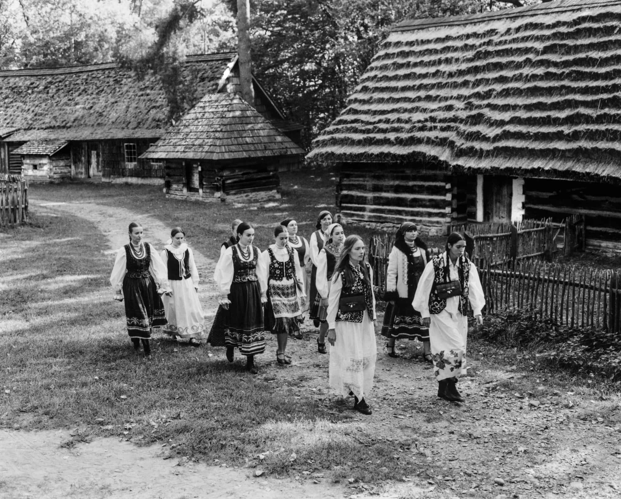 Black and white image of a group of women in traditional folk costumes walking along a dirt path between rustic thatched-roof wooden cottages.