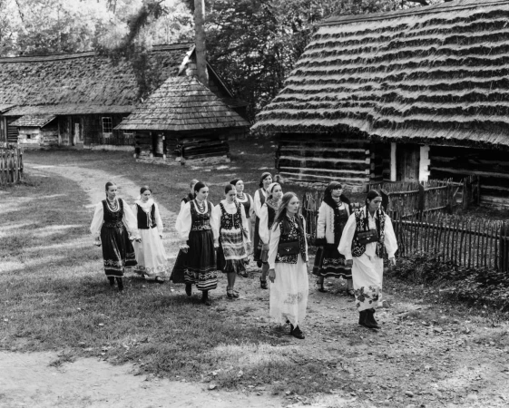 Black and white image of a group of women in traditional folk costumes walking along a dirt path between rustic thatched-roof wooden cottages.