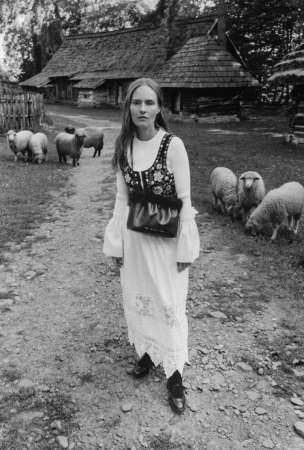 Model wearing a white dress and patterned vest holding a black handbag outdoors with grazing sheep and a rustic cottage background