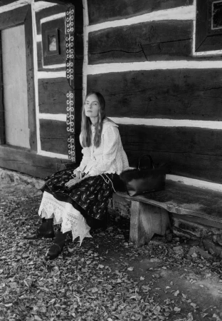 Model seated on a wooden bench against a rustic log cabin wall for Chylak fall 2024 campaign, captured in black and white by Stanisław Boniecki