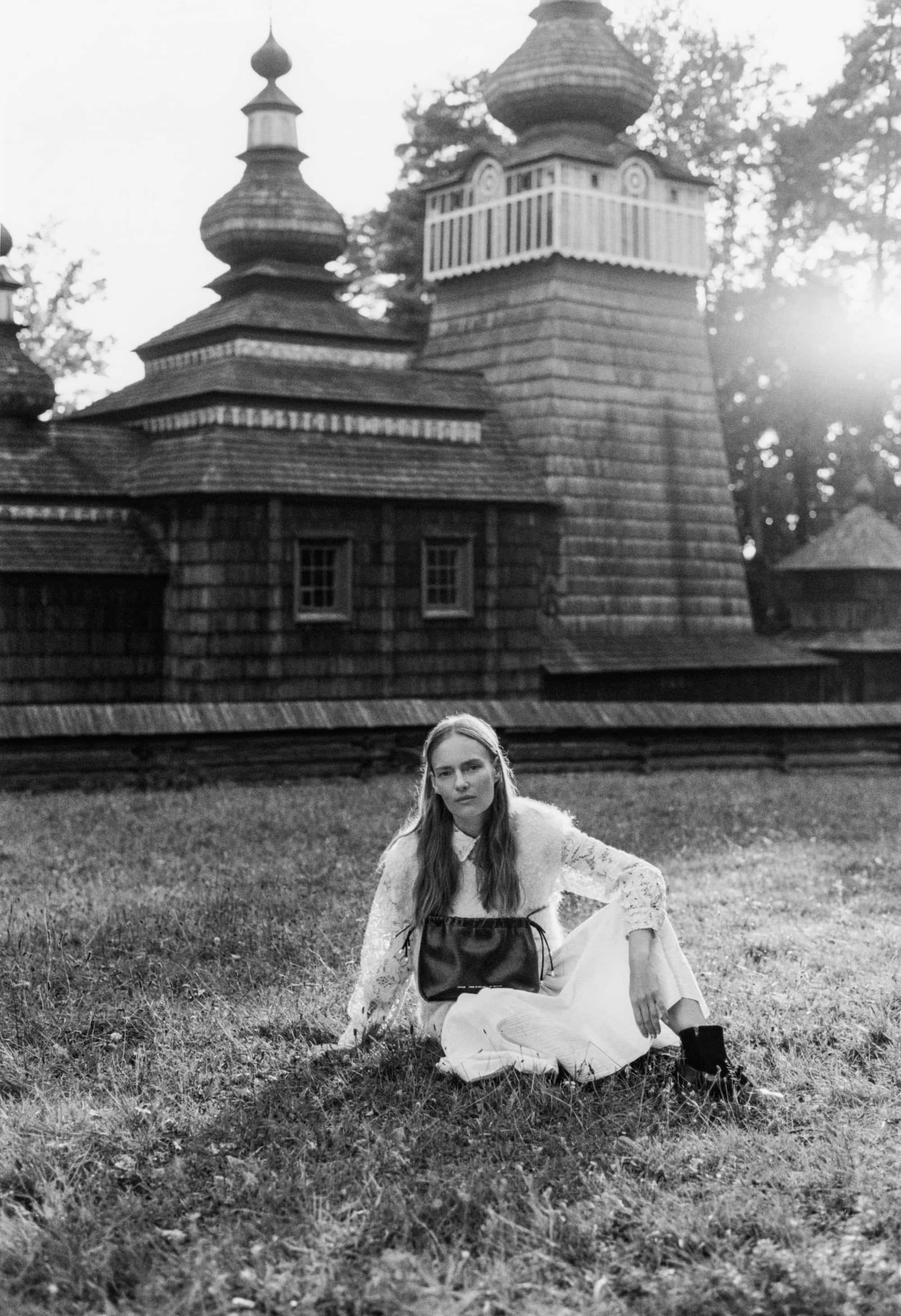 Model seated on grass holding a leather handbag in front of a wooden church with onion domes for Chylak.