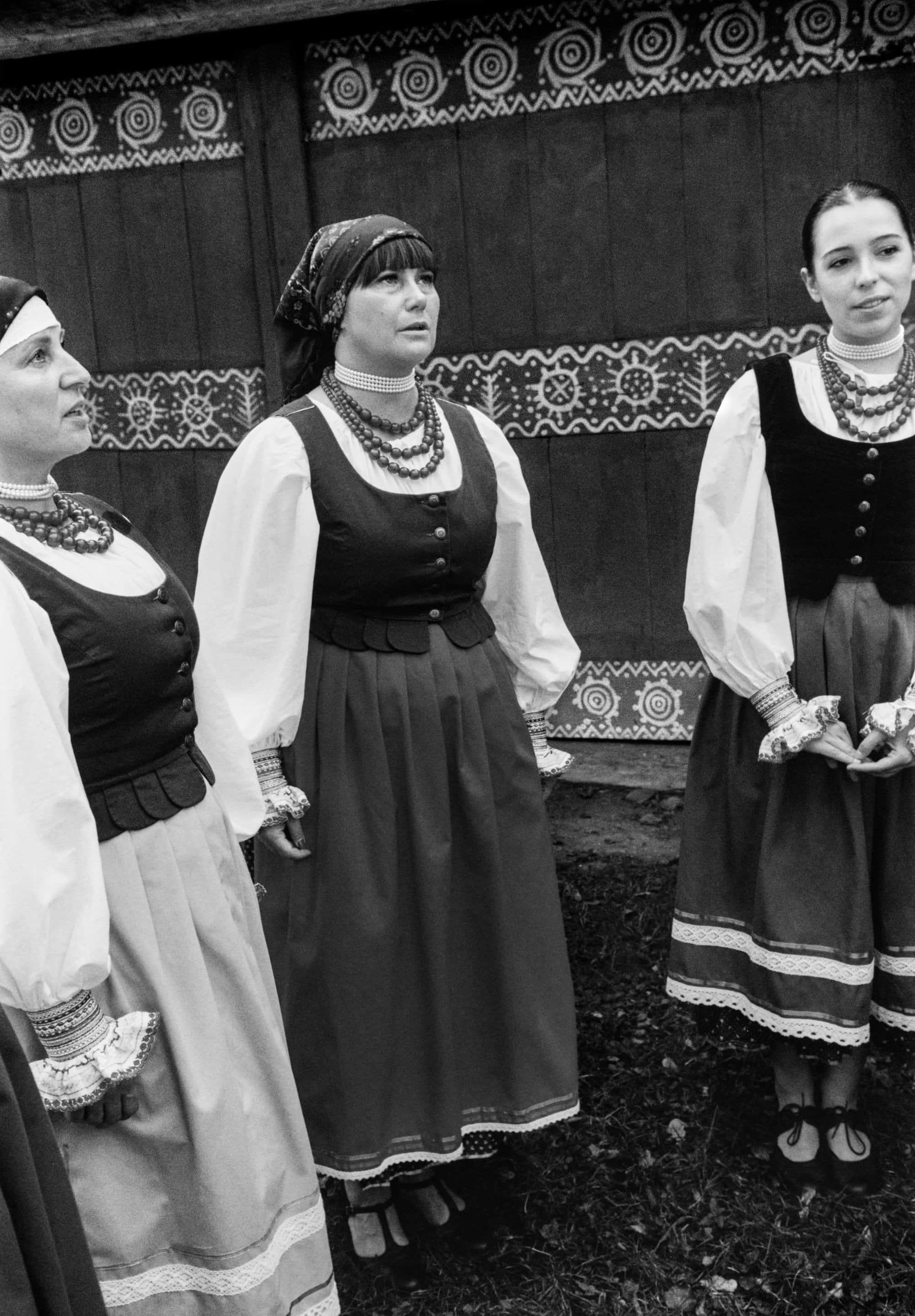 Three women in traditional folk costumes standing outdoors against a decorated wooden wall in a black and white photograph