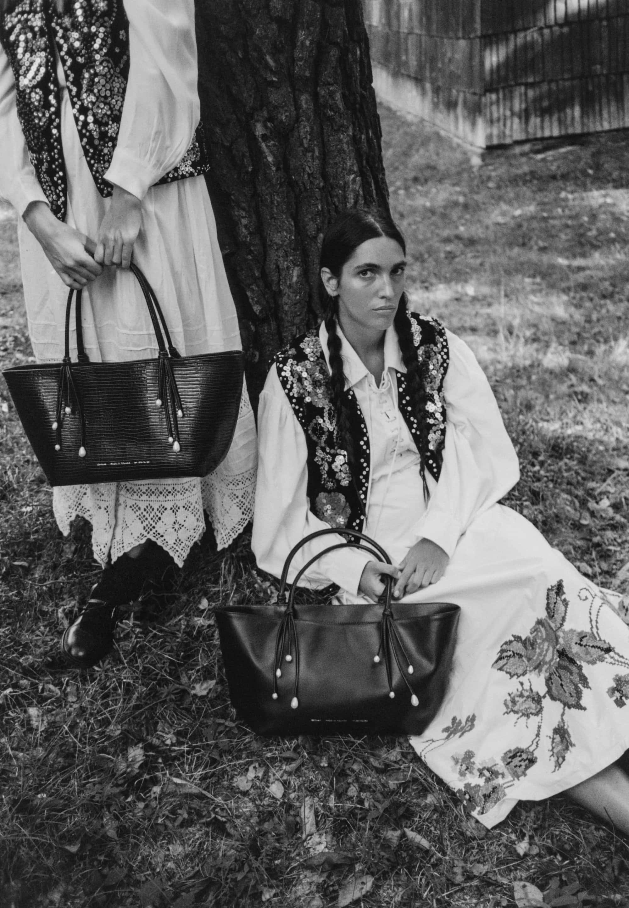 Two women in white dresses and embroidered vests seated outdoors holding black leather tote bags against a tree trunk.