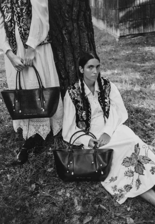 Two women in white dresses and embroidered vests seated outdoors holding black leather tote bags against a tree trunk.