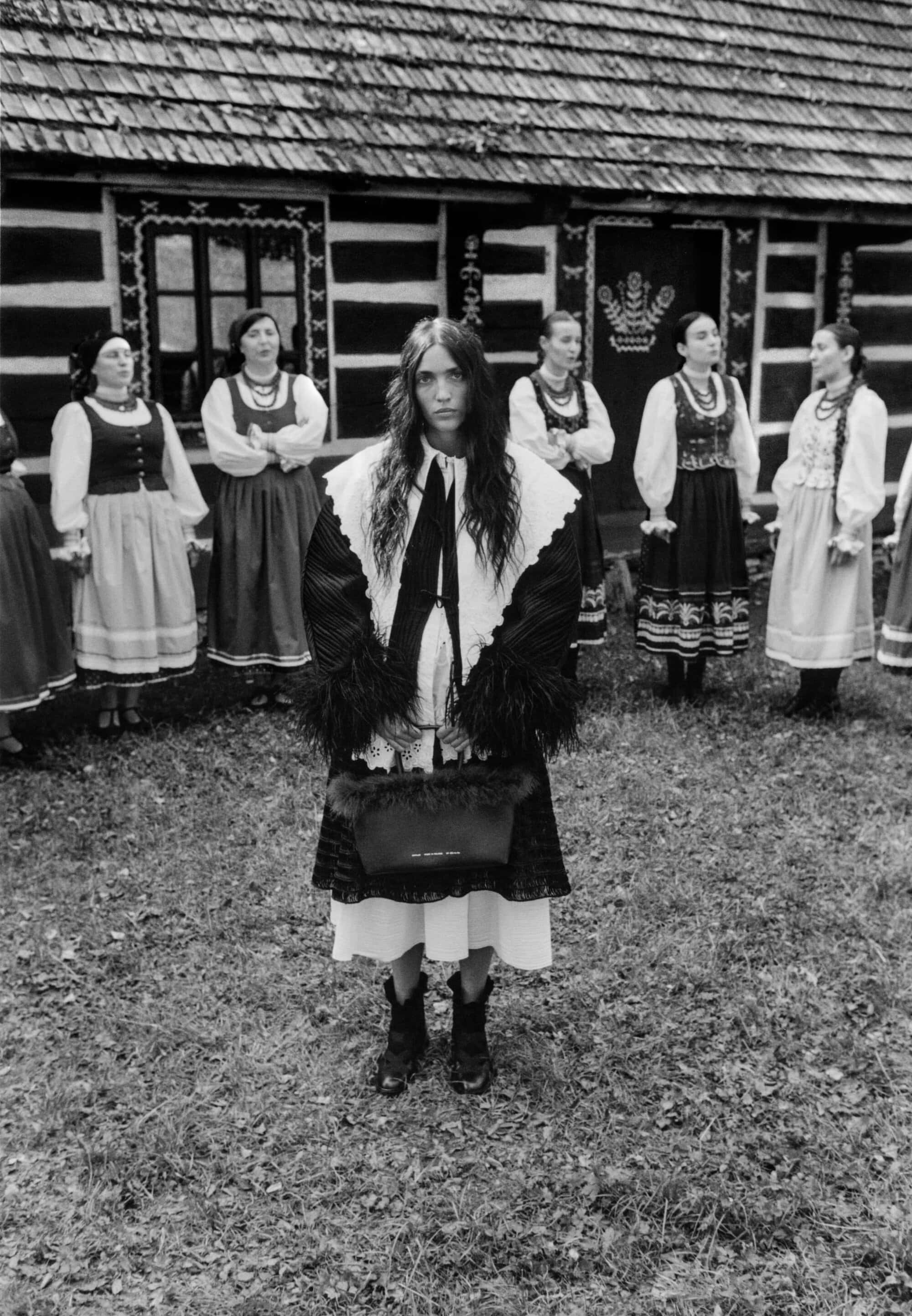 A woman holding a dark handbag stands centrally in foreground, with a group of women in traditional folk costumes standing in background.
