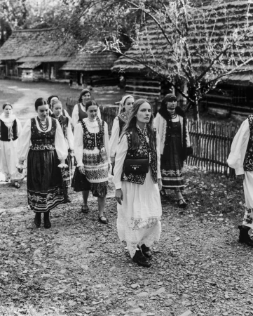 Group of women in traditional folk costumes walking along a path past rustic wooden cottages.