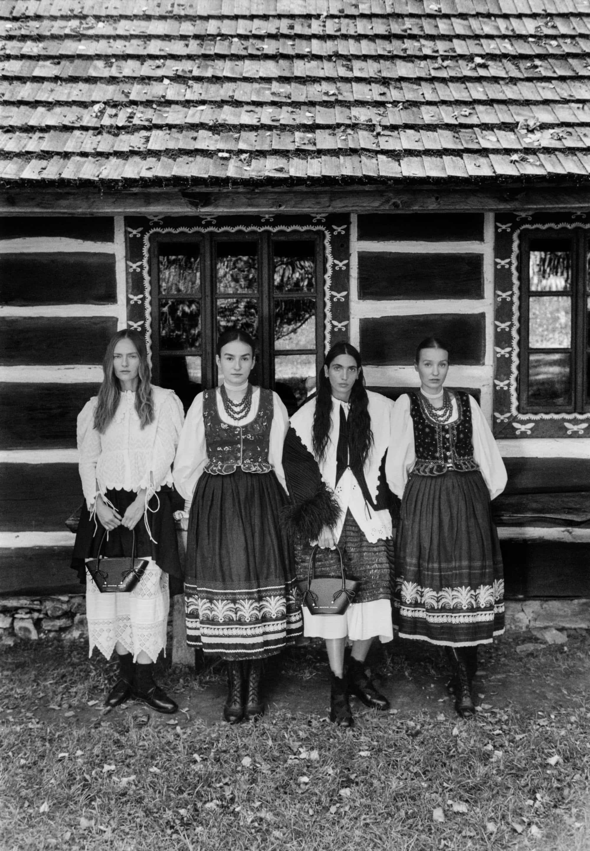 Four female models standing in traditional attire holding handbags in front of a rustic wooden building exterior.