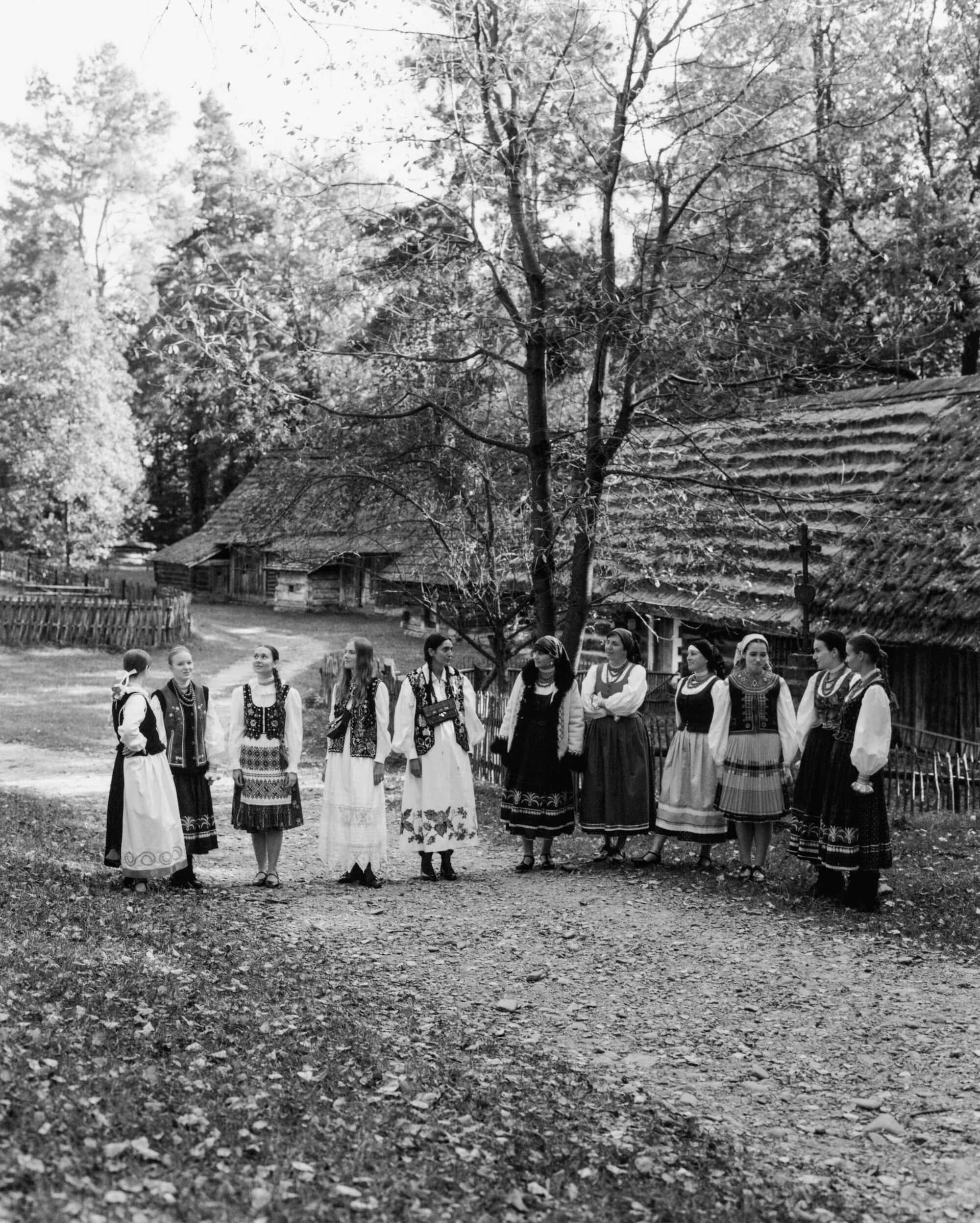 Group of women in traditional folk costumes standing along a dirt path near thatched-roof wooden cottages