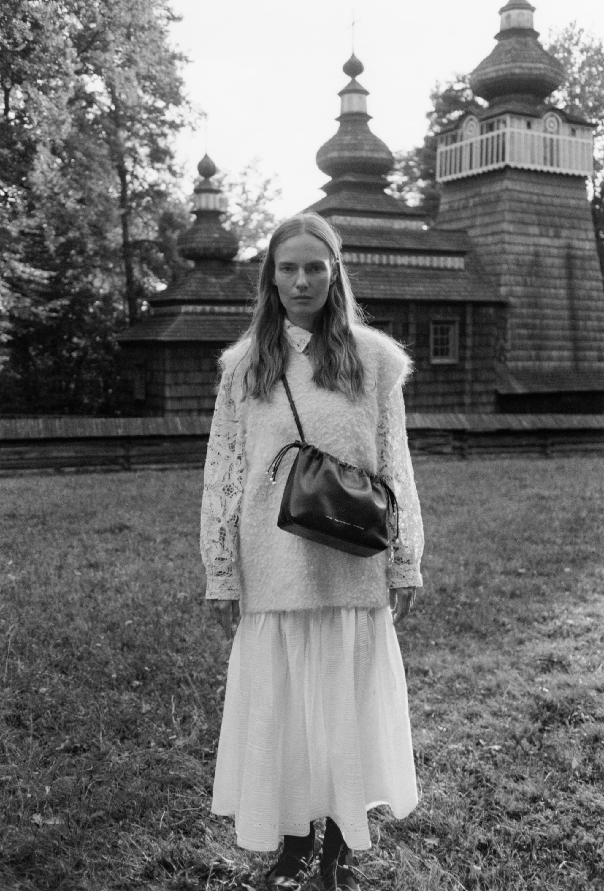 Monochrome fashion campaign image featuring a model wearing a textured sweater and long skirt carrying a dark leather shoulder bag in front of a wooden church.