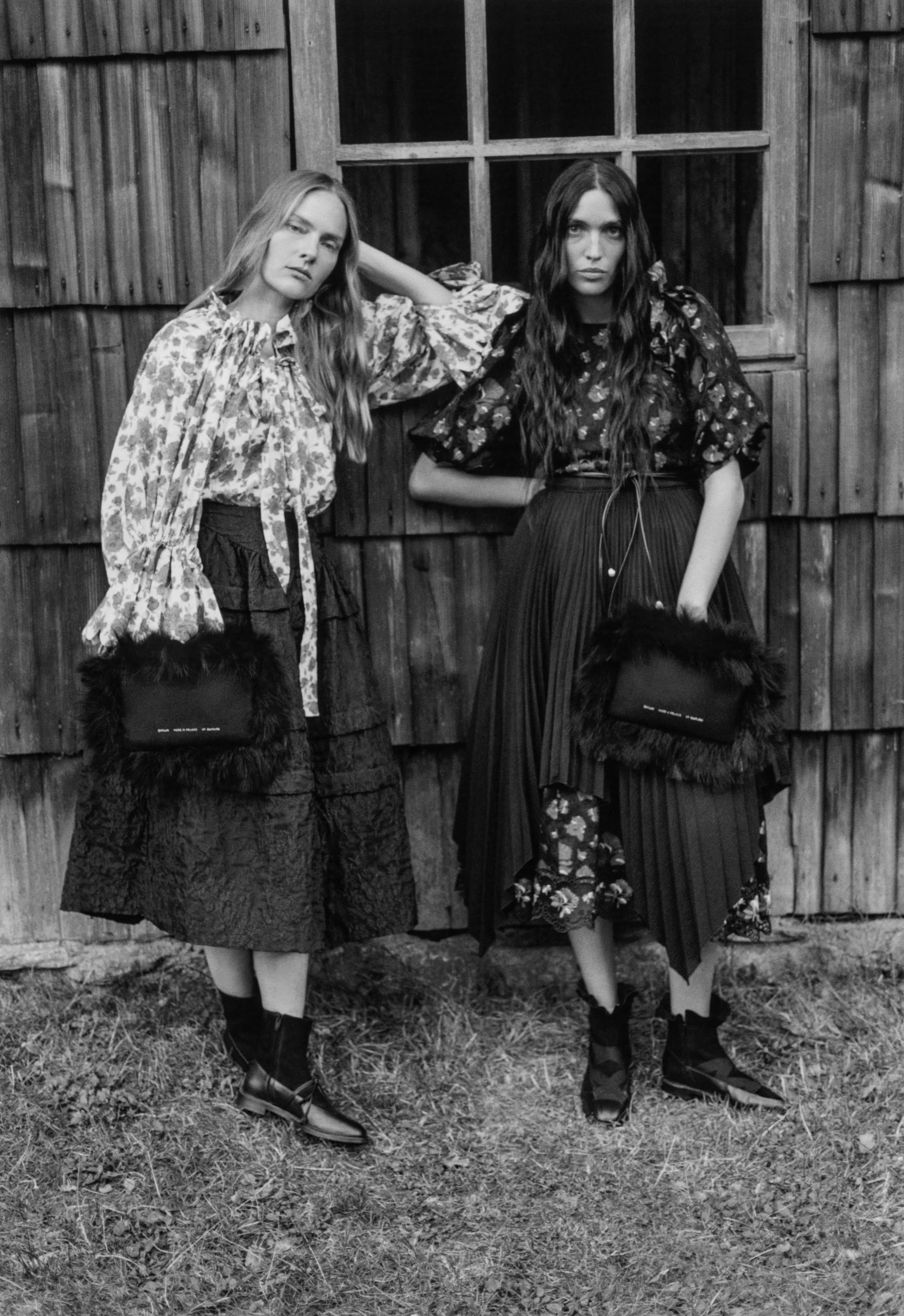 Two female models in patterned tops and dark skirts posing outdoors against a rustic wooden wall holding furry handbags.