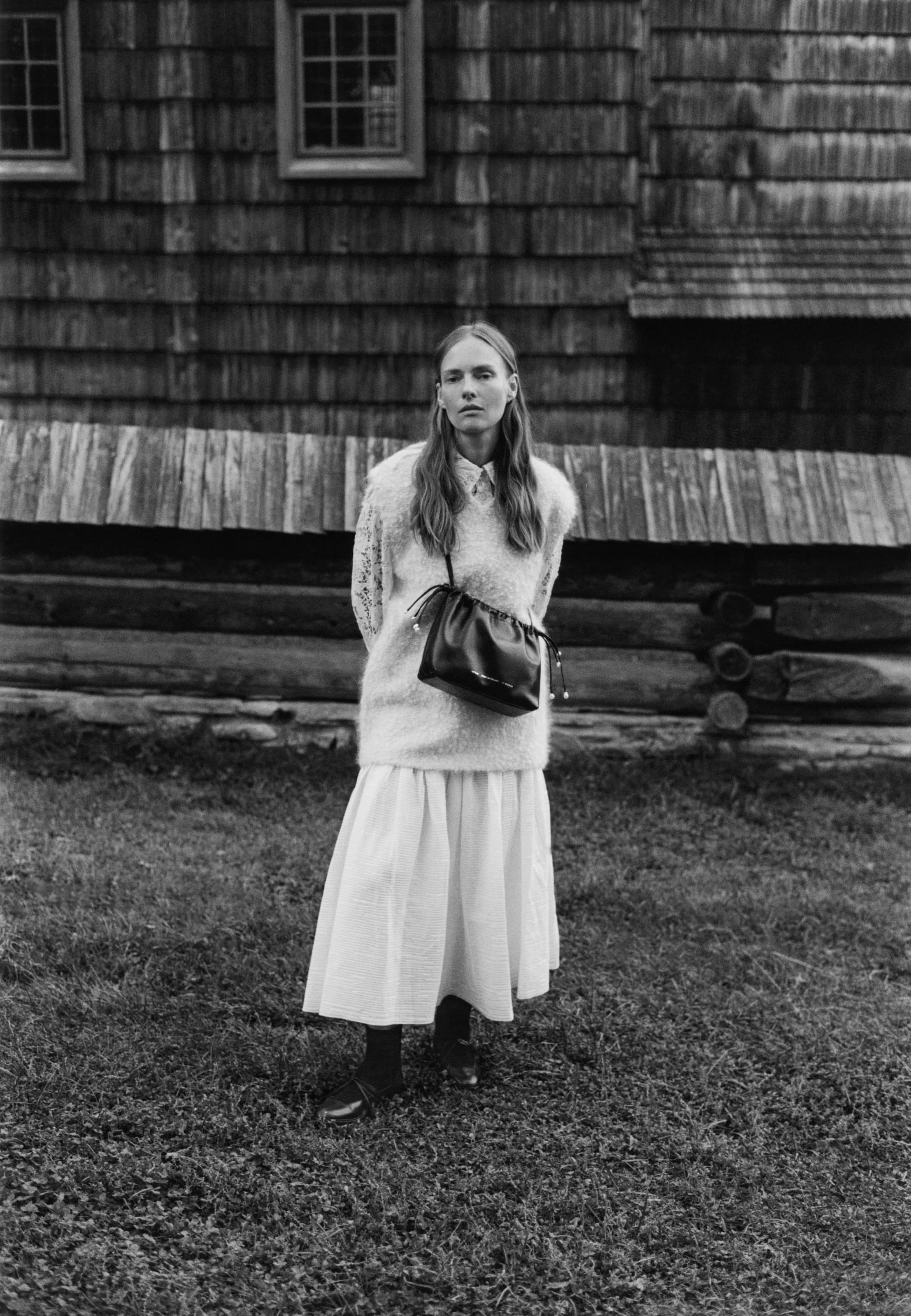 Model standing outdoors wearing a textured knit sweater and long skirt with a leather handbag, in front of a wooden building.