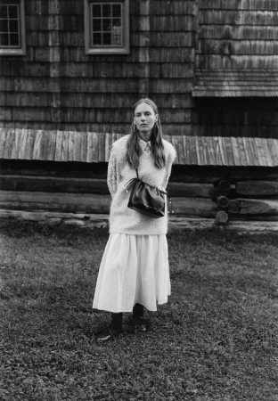 Model standing outdoors wearing a textured knit sweater and long skirt with a leather handbag, in front of a wooden building.