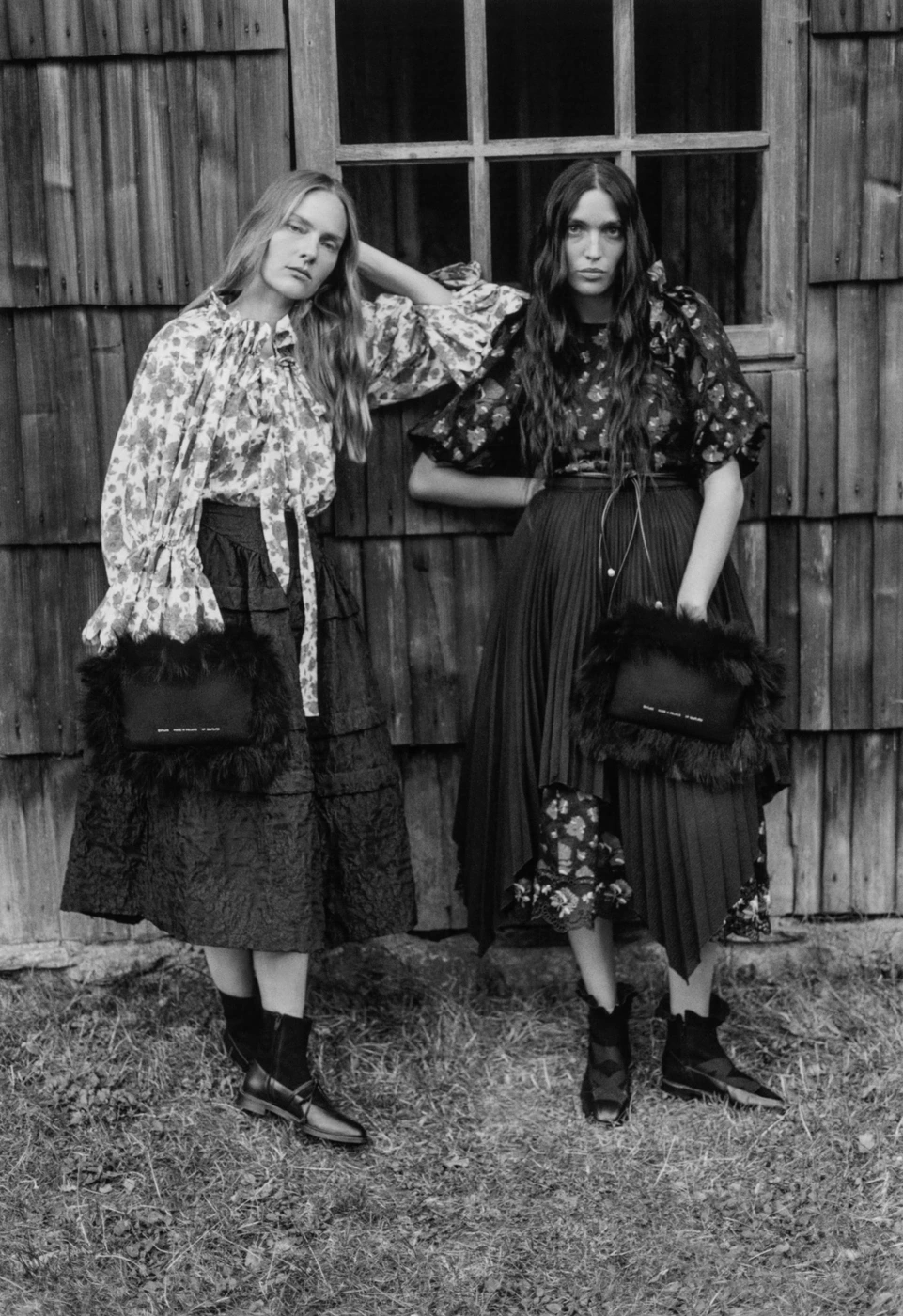 Two female models in patterned tops and dark skirts posing outdoors against a rustic wooden wall holding furry handbags.