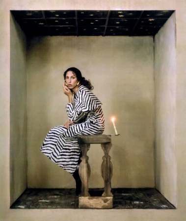 An adult woman in a striped dress sits on a wooden stool next to a lit candle in a tan colored room with a decorative ceiling.