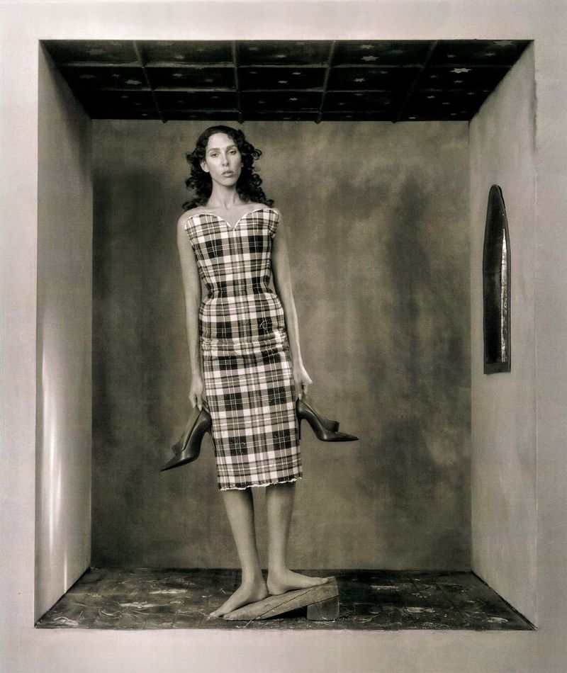A woman in a plaid dress poses barefoot in a box-like room, holding high heels. The room has textured walls and a patterned ceiling.