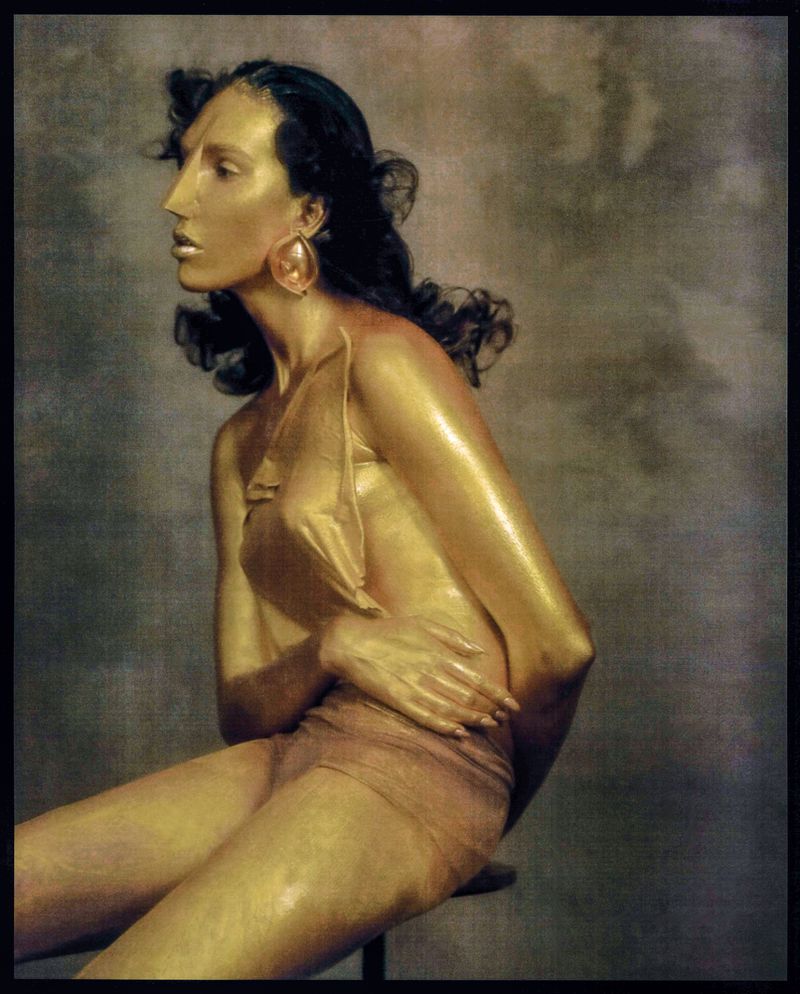 Woman with gold body paint sits in profile wearing a dress, large earrings, and dark wavy hair against a mottled gray background.