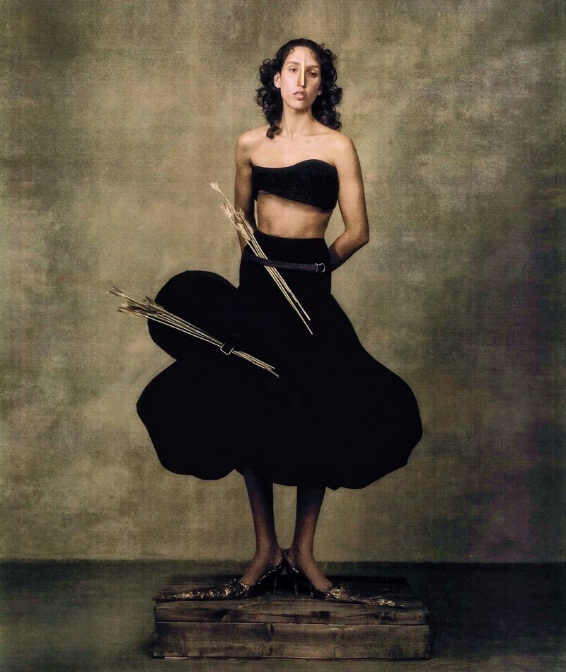 A fashion portrait of a slender woman wearing a black two-piece outfit with unique shoes, standing on a wooden crate against a painted backdrop.